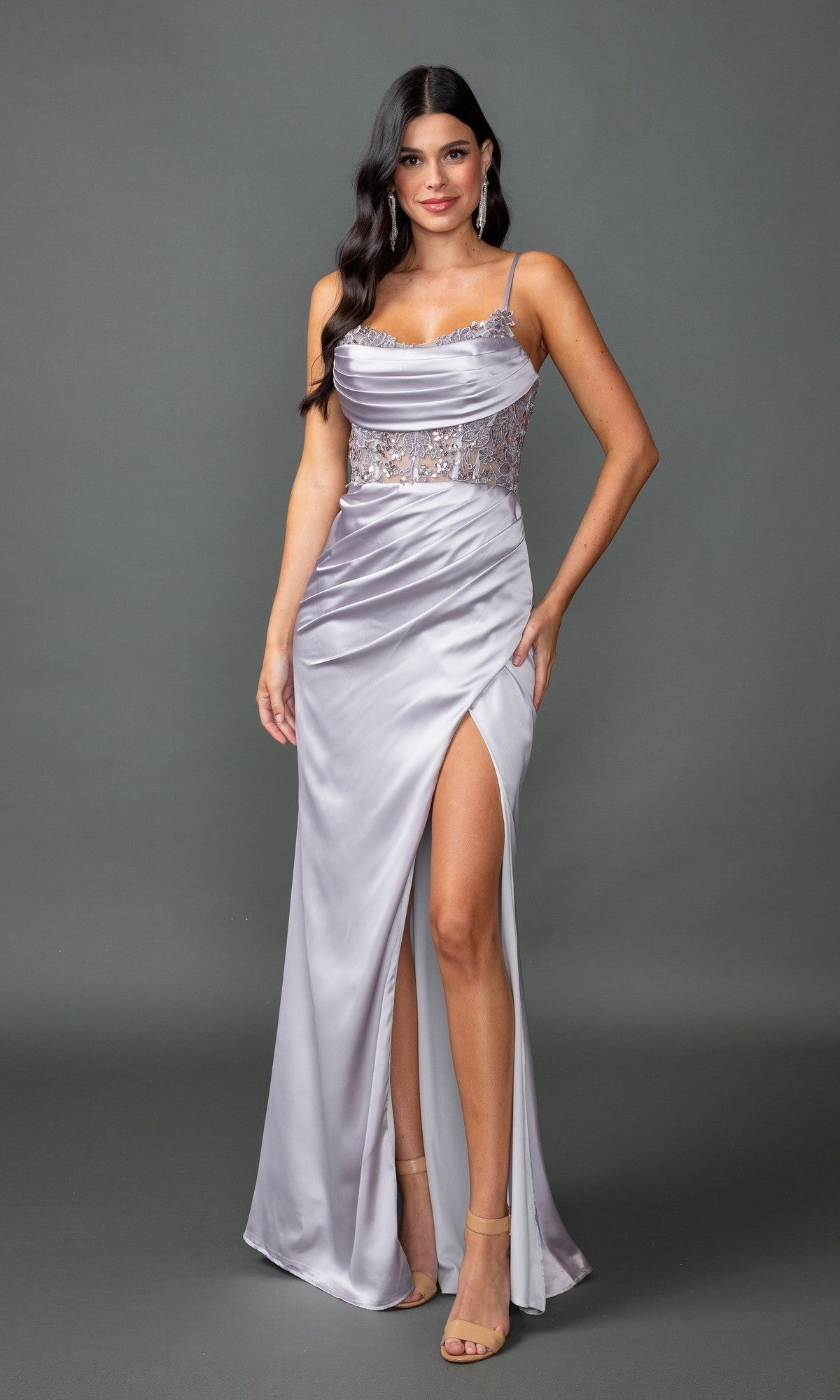 A woman poses against a plain gray background in the Sheer-Waist Long Prom Dress: Dancing Queen 4416, a floor-length silver satin gown with a thigh-high slit, embellished bodice, and spaghetti straps.